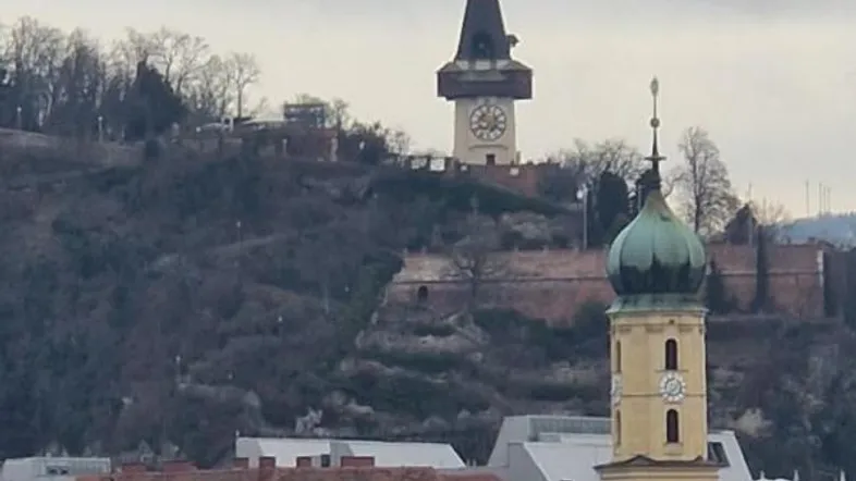 Stadtansicht mit dem Grazer Uhrturm und Dächern im Vordergrund unter bewölktem Himmel.