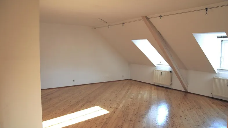 Helles Dachzimmer mit schrägen Wänden, Holzboden, sichtbarem Balken und Dachfenstern, ideal als Schlafzimmer.