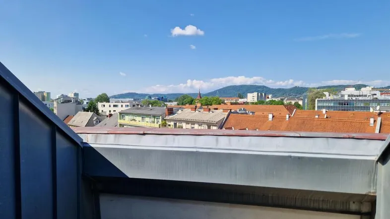 Blick von einem Dachfenster über die Dächer der Stadt mit Bergen im Hintergrund an einem sonnigen Tag.