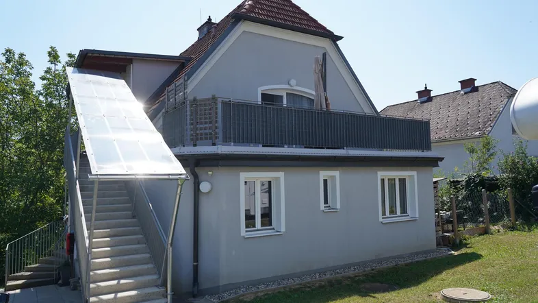Modernes Gebäude mit grauer Fassade, Balkon und einer Außentreppe mit Rampe, umgeben von Grün.