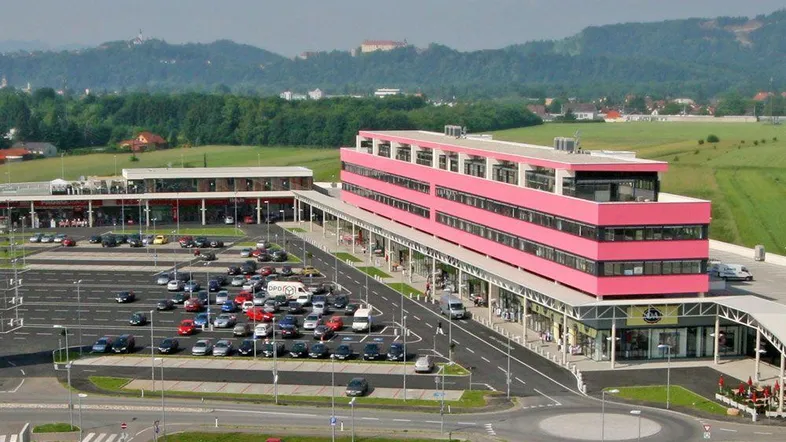 Luftaufnahme eines modernen Einkaufszentrums mit großem Parkplatz und grüner Hügellandschaft im Hintergrund.