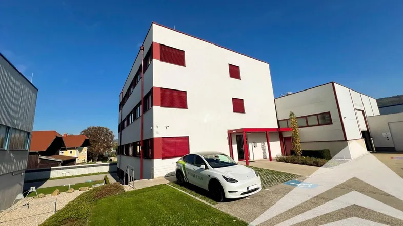 Modernes Bürogebäude mit weißer Fassade, roten Fensterläden und einem gepflegten Außenbereich unter blauem Himmel.