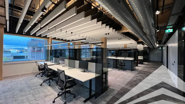 Moderner Großraumbüro-Arbeitsbereich mit ergonomischen Stühlen, großen Fenstern und sichtbaren Lüftungsrohren.