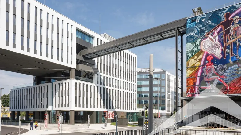 Moderne Bürogebäude mit markanter Architektur, einer Verbindungsbrücke und einem großen farbenfrohen Wandgemälde.