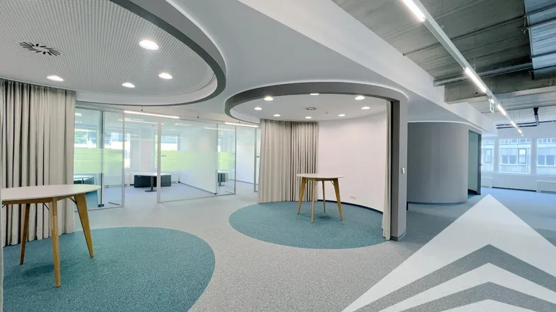 Moderner Bürobereich mit Teppichboden in Grau und Türkis, Glaswänden und indirekter Beleuchtung.