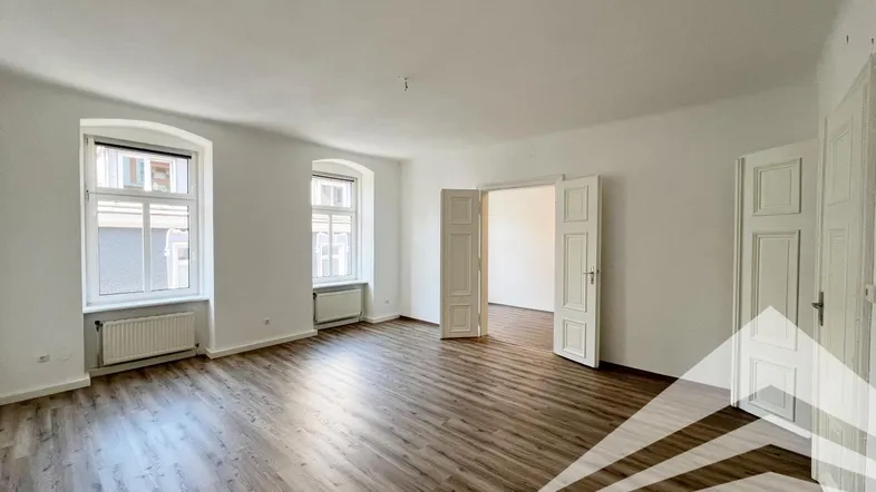 Heller Wohnraum mit Laminatboden, hohen Fenstern und Zugang zu einem weiteren Zimmer.