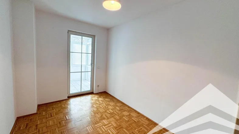 Helles Zimmer mit Parkettboden und Zugang zu einem verglasten Balkon oder einer Loggia.