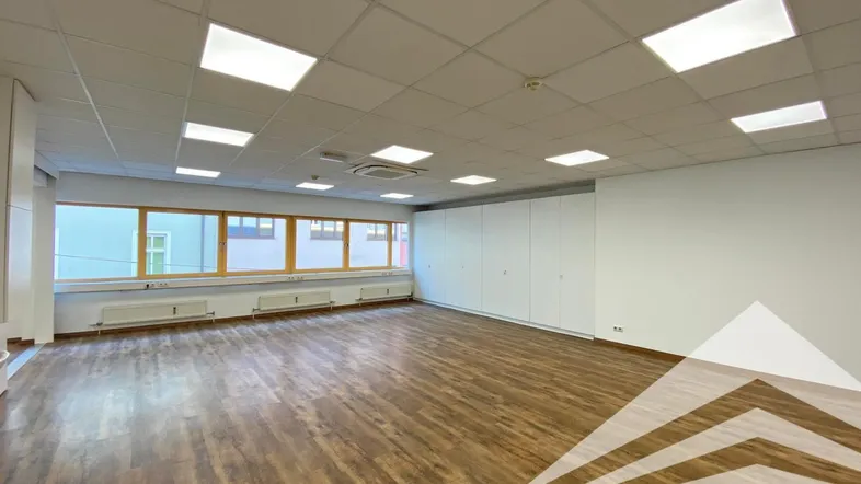 Geräumiges Büro mit Holzfußboden, großen Fenstern und abgehängter Decke mit integrierter Beleuchtung.