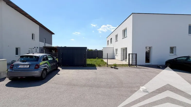 Moderne Neubauten mit Parkplätzen und kleiner Grünfläche unter blauem Himmel.