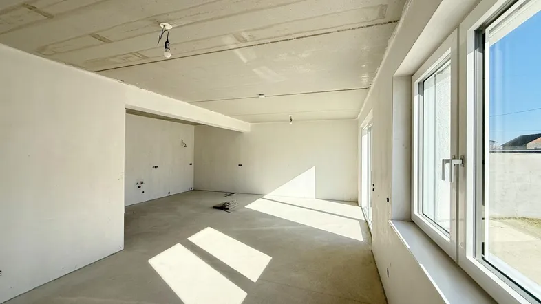 Heller, unmöblierter Wohnbereich im Rohbau mit großen Fenstern und viel Tageslicht.