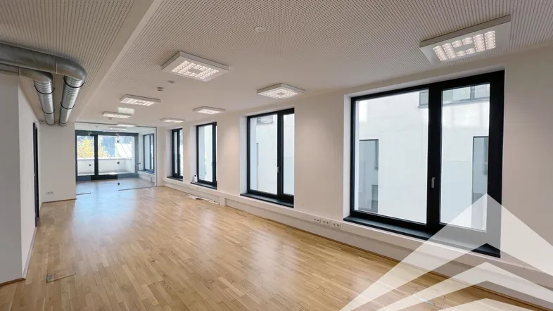 Geräumiger, leerer Büroraum mit Holzboden, großen Fenstern und Zugang zu einem Balkon oder einer Terrasse.