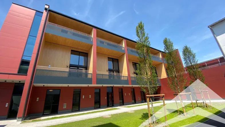Moderne Wohnanlage mit roter Fassade, Holzverkleidung und begrüntem Innenhof unter blauem Himmel.