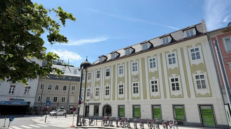 Historisches Gebäude an einer Straßenecke mit vielen Fenstern und Geschäften im Erdgeschoss unter blauem Himmel.