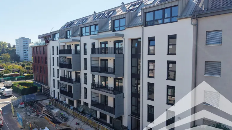 Modernes Mehrfamilienhaus im Bau mit Balkonen und Dachgauben, umgeben von einer Baustelle.
