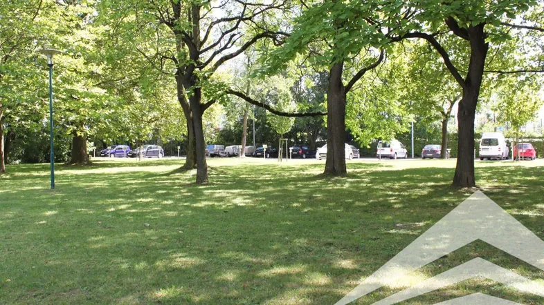 Grüner Parkplatzbereich mit Bäumen und gepflegtem Rasen, im Hintergrund sind parkende Autos zu sehen.
