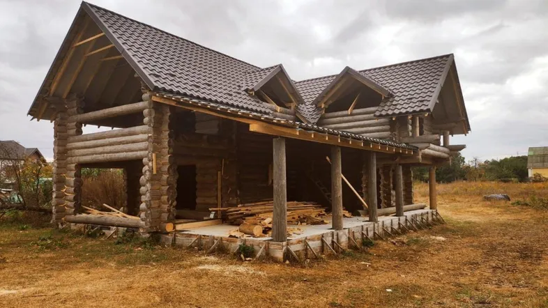 Holzblockhaus im Bau mit sichtbarer Holzkonstruktion und Dachdeckung unter bewölktem Himmel.