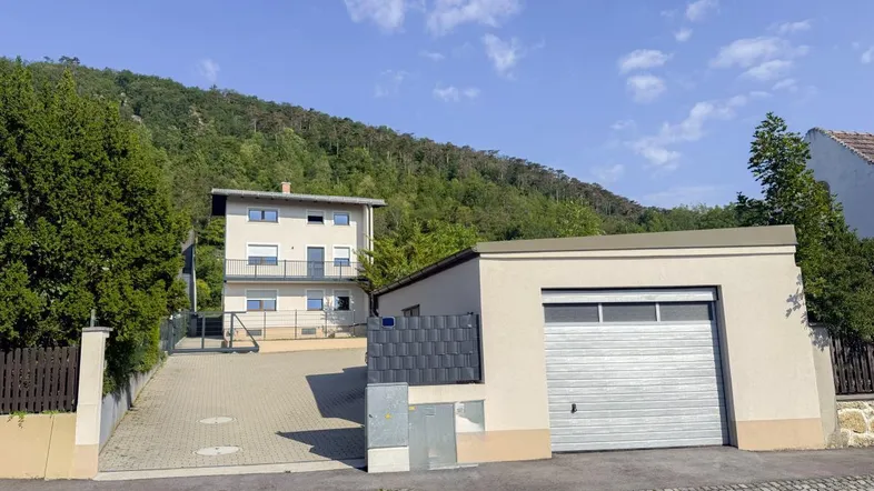 Hausfassade mit Garage und Zufahrt, umgeben von grünen Hügeln unter blauem Himmel.