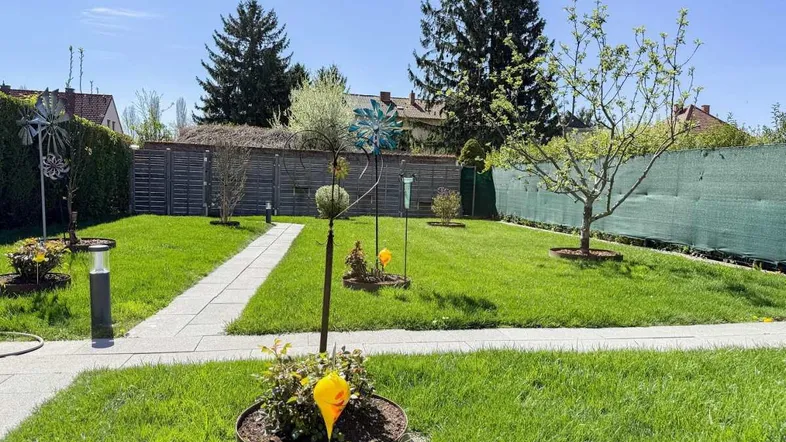 Gepflegter Garten mit Rasenfläche, Gehwegen und dekorativen Elementen unter blauem Himmel.