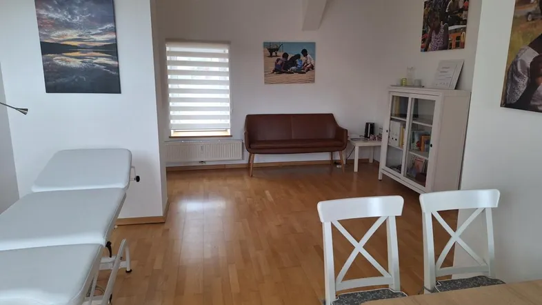 Heller Büroraum mit Holzboden, einer Untersuchungsliege und einem Wartebereich mit Sofa und Stühlen.