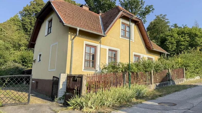 Gelbes Einfamilienhaus mit rotem Ziegeldach und Holzzaun in ländlicher Umgebung.