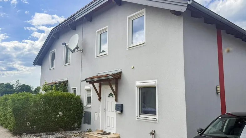 Graue Hausfassade mit weißen Fenstern, Haustür und Satellitenschüssel unter bewölktem Himmel.