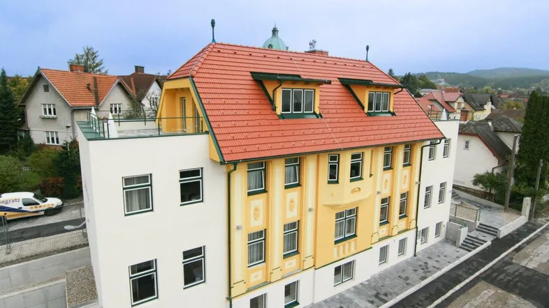 Gebäudeansicht von oben mit rotem Ziegeldach und gelber Fassade, umgeben von weiteren Häusern.