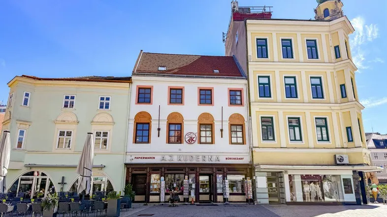Historische Gebäude an einem sonnigen Stadtplatz mit Geschäften und Außengastronomie.