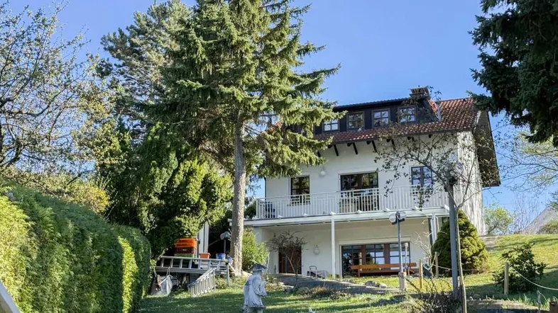 Freistehendes Haus mit weißer Fassade, Balkonen und einem gepflegten Garten mit hohen Bäumen.