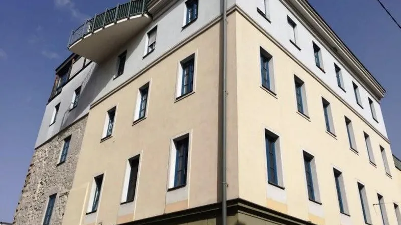 Mehrstöckiges Gebäude mit heller Fassade, blauen Fenstern und einem kleinen Balkon.