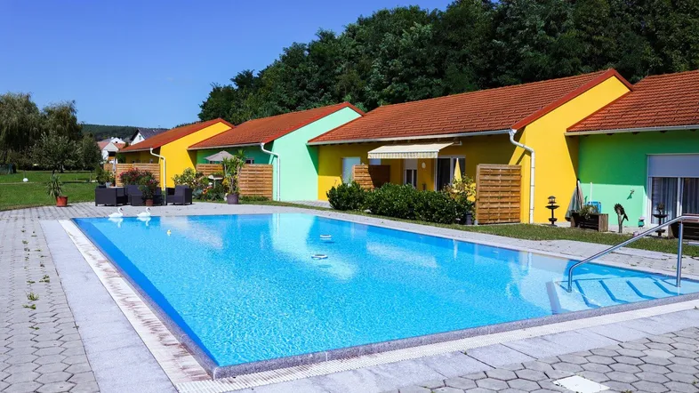 Großer Außenpool umgeben von bunten Häusern und einer gepflegten Grünfläche unter blauem Himmel.