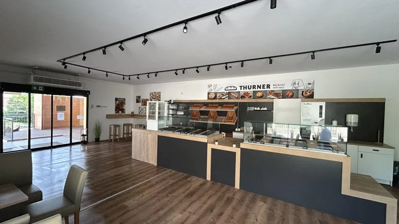 Moderner Verkaufsraum mit Holzboden, Theken und Sitzgelegenheiten, ideal für ein Café oder eine Bäckerei.