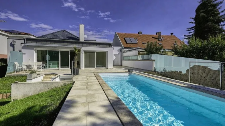 Modernes Haus mit langem Swimmingpool, gepflasterter Terrasse und Gartenbereich unter blauem Himmel.