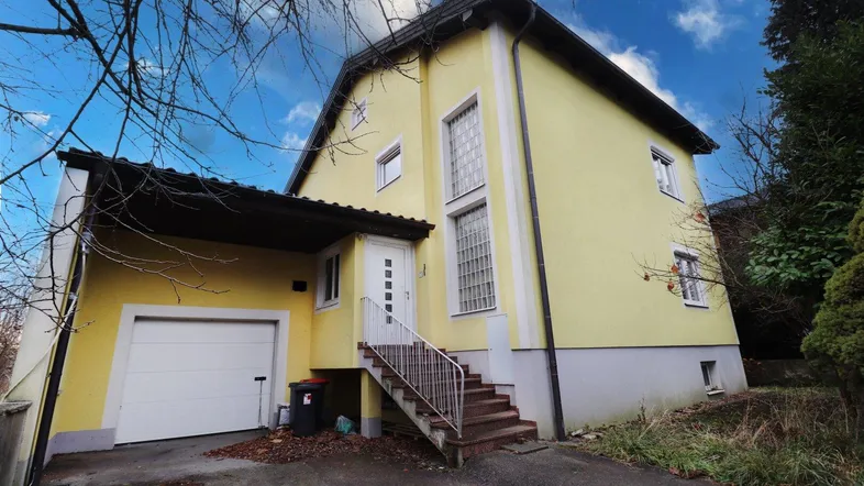 Gelbes Einfamilienhaus mit Garage, Außentreppe und Carport unter bewölktem Himmel.