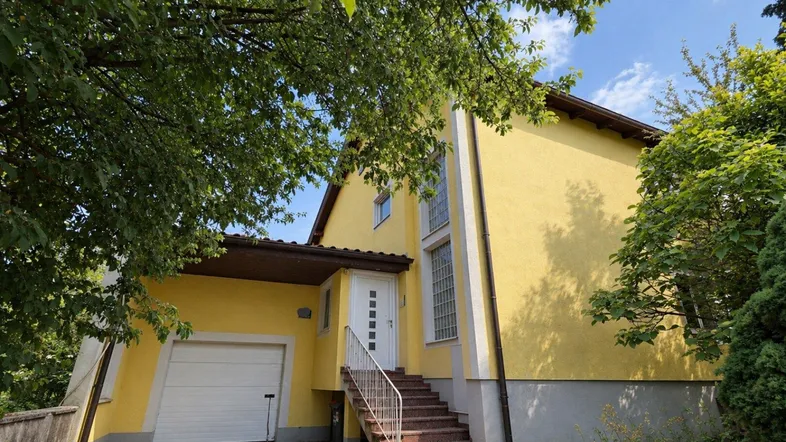 Gelbes Haus mit weißer Eingangstür und integrierter Garage, umgeben von Bäumen und einem gepflasterten Weg.