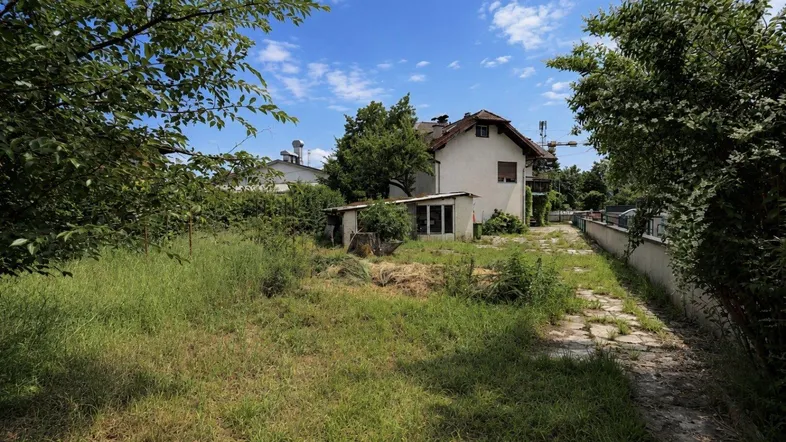 Verwilderter Gartenbereich mit altem Schuppen und einem Wohnhaus im Hintergrund unter blauem Himmel.
