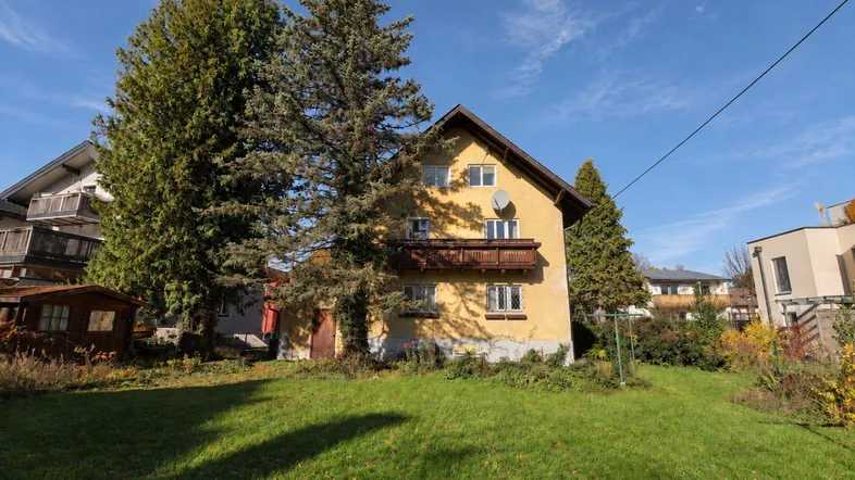 Gelbes Wohnhaus mit Holzbalkon und großem Garten, umgeben von hohen Bäumen unter blauem Himmel.