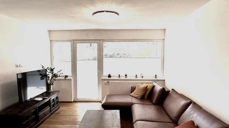 Gemütliches Wohnzimmer mit braunem Ecksofa, Holzboden und Zugang zu einem Außenbereich.