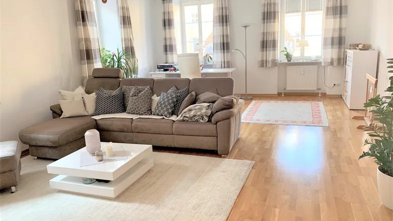 Helles Wohnzimmer mit bequemer L-förmiger Couch, Holzboden und mehreren Fenstern für viel Tageslicht.