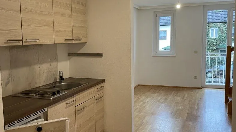 Helle offene Wohnfläche mit integrierter Küchenzeile, Parkettboden und Zugang zu einem Balkon.