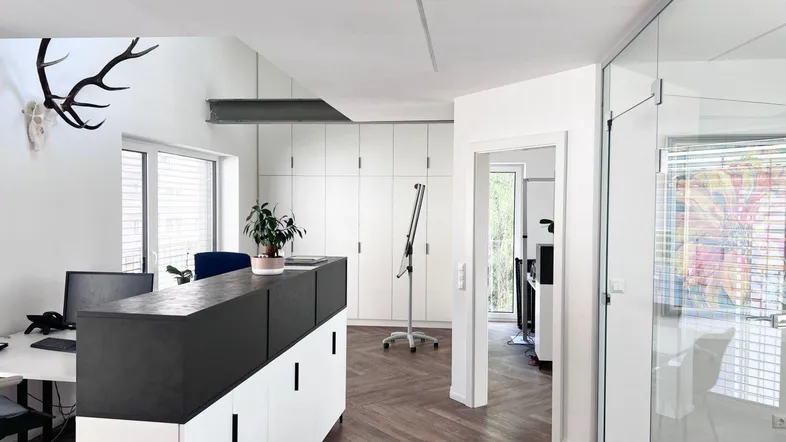 Modernes Büro mit Fischgrätparkett, weißen Einbauschränken und viel Tageslicht durch große Fenster.