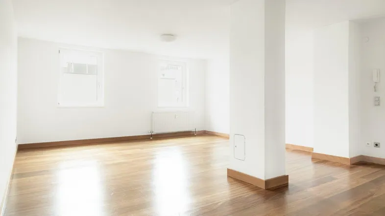 Heller, leerer Wohnraum mit glänzendem Parkettboden, weißen Wänden und zwei Fenstern.