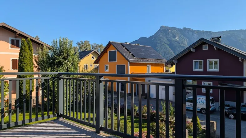 Balkon mit Holzboden und schwarzem Geländer bietet einen weiten Blick auf die umliegende Berglandschaft und Nachbarhäuser.