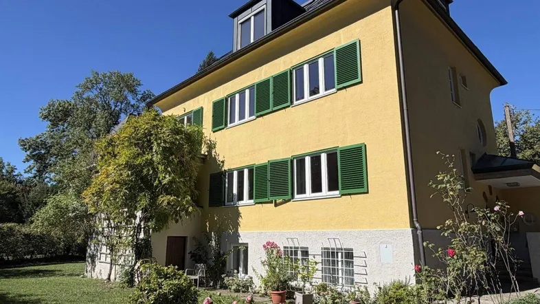 Gelbes Haus mit grünen Fensterläden und gepflegtem Vorgarten unter blauem Himmel.