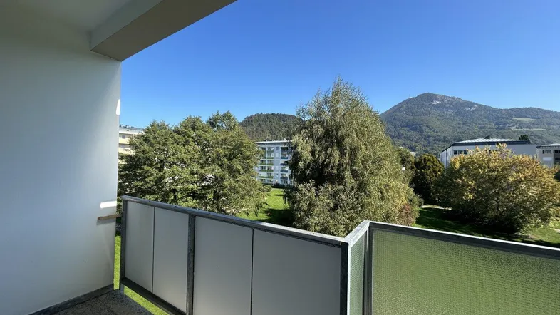 Heller Balkon mit Blick auf grüne Bäume und bewaldete Berge unter blauem Himmel.