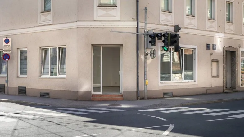 Eckgebäude mit leerstehendem Ladenlokal im Erdgeschoss an einer belebten Straßenkreuzung mit Ampel und Zebrastreifen.