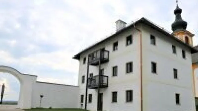 Weißes Gebäude mit schwarzen Balkonen und einem Kirchturm im Hintergrund unter bewölktem Himmel.