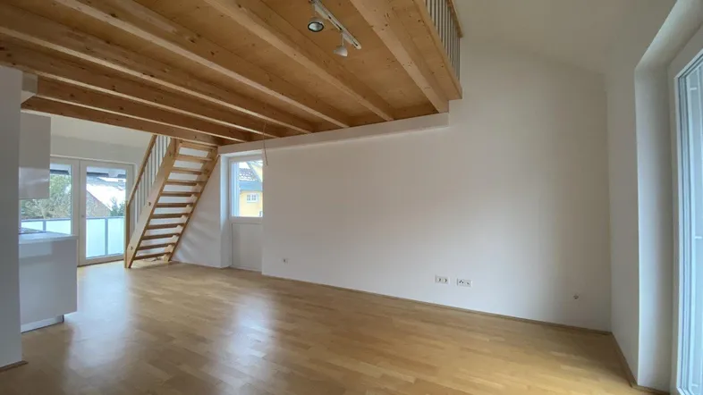 Heller Wohnbereich mit Holzboden, sichtbaren Deckenbalken und einer Holztreppe zum oberen Geschoss.