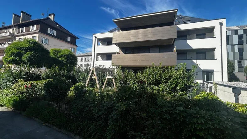 Blick auf ein modernes Gebäude mit Balkonen und ein älteres Haus, umgeben von einem grünen Garten mit Schaukel.