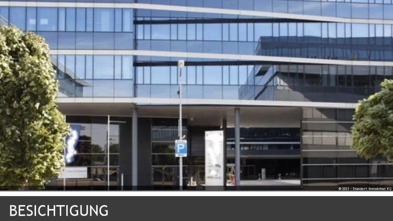 Moderne Bürogebäude-Fassade mit großen Glasflächen und reflektierenden Fenstern, umgeben von grünen Bäumen.