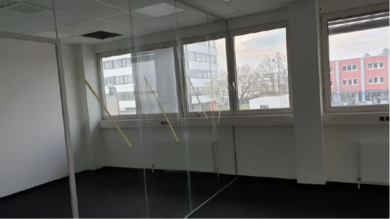 Heller Büroraum mit großen Fenstern und Blick auf umliegende Gebäude, ausgestattet mit Teppichboden.